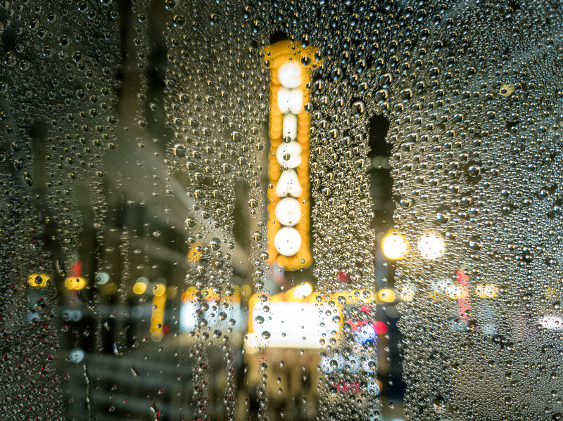 Rain-covered window with blurred city lights and an illuminated tall building in the background.