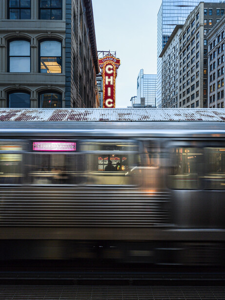 Motion-blurred subway train passing beneath vintage theater marquee and modern downtown buildings.