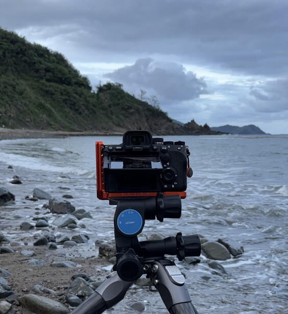 Camera mounted on tripod on rocky beach with ocean and cloudy sky in background.