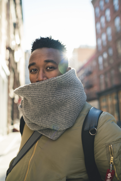 Person wearing olive jacket and gray knit neck gaiter on urban street with buildings behind.