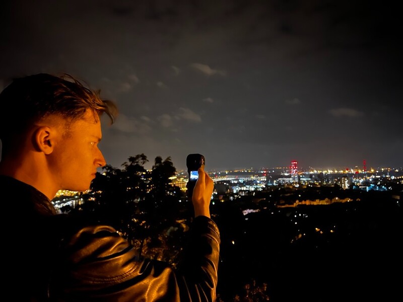 Man holding smartphone capturing nighttime cityscape from elevated vantage point.
