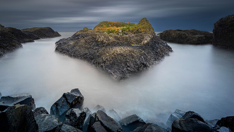 Seascape with dark rocky outcrops surrounded by smooth, misty water under a moody sky.