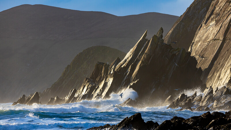 Dramatic coastal cliffs with columnar basalt formations rise above turbulent ocean waves under clear sky.