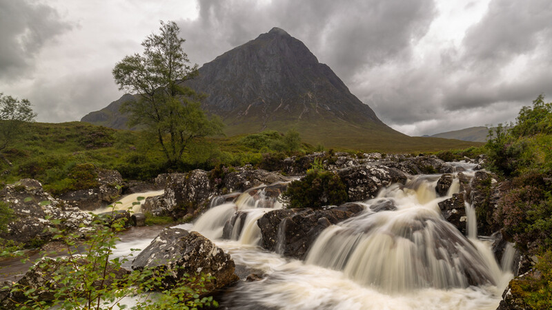 Rapid river cascading over dark rocks beneath a steep conical mountain under stormy skies.