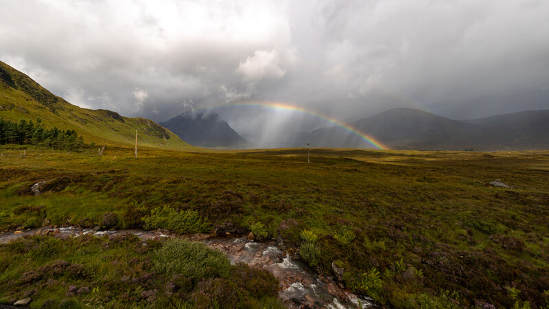 Rainbow extending from storm clouds over a vast green valley with mountains in the background.