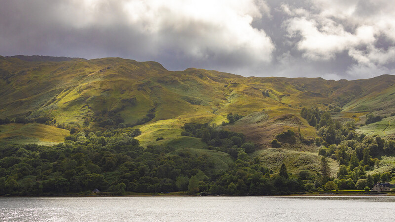 Rolling green hills reflected in still water, with forested valleys and dramatic cloudy sky.