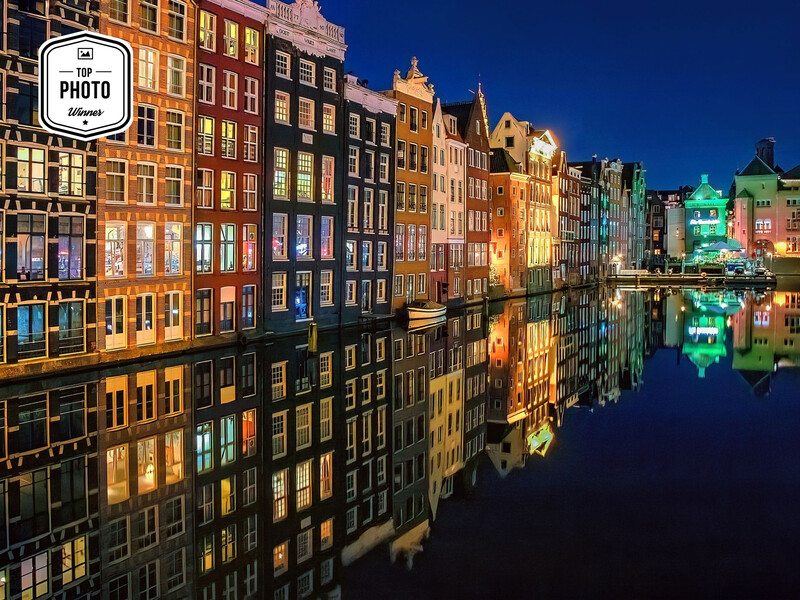 Illuminated canal-side buildings reflected in still water at dusk, with warm and cool-toned architectural lighting.