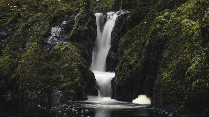 Waterfall cascading through moss-covered rock formations in a narrow canyon.