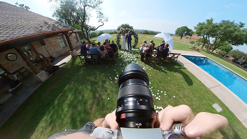 First-person view holding a telephoto lens at an outdoor gathering near a swimming pool.