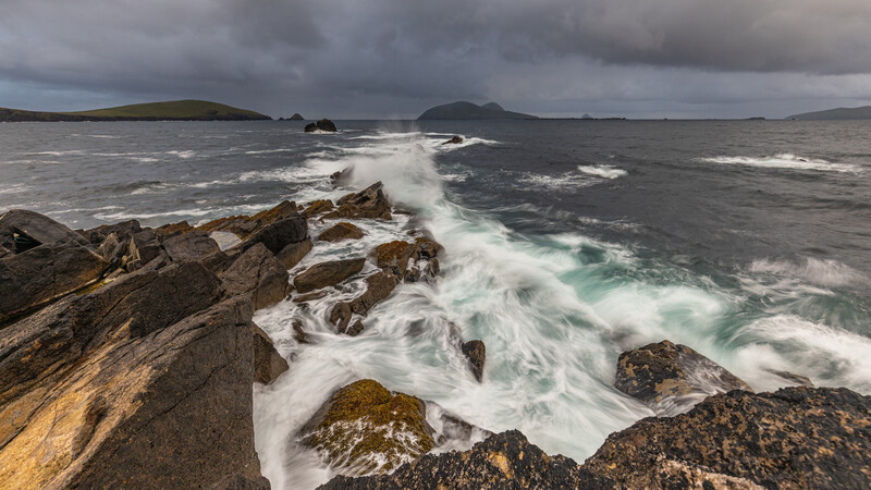 Waves crash against rocky shoreline with mountains visible across the bay under stormy skies.