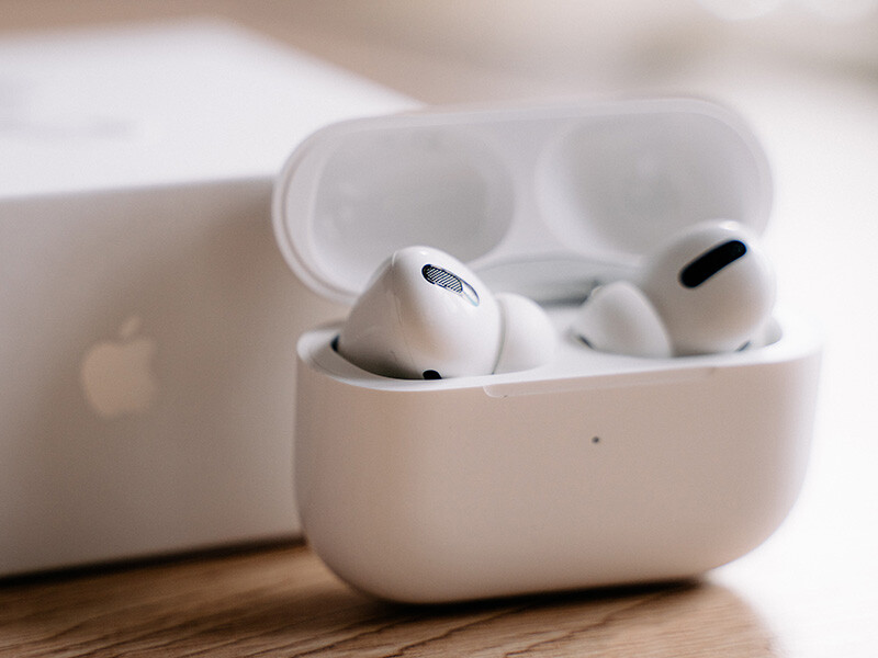 White wireless earbuds in an open charging case next to a laptop, shot in natural daylight.