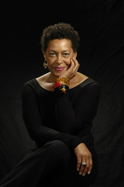 Woman in black long-sleeve top poses against dark background with hand near face.
