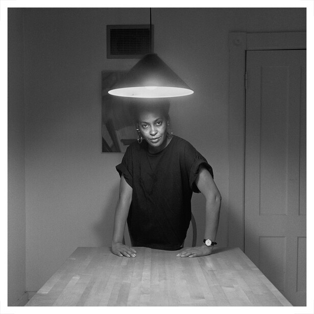 Person leaning on table under dramatic overhead pendant light in black and white studio setting.