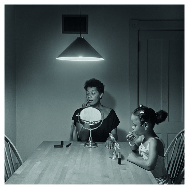 Two women at a table under a pendant light, one holding a mirror while the other applies makeup.