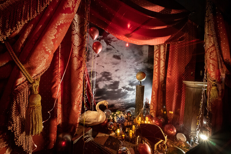 Elaborate still life altar arrangement with red draped fabric, celestial imagery, and ornamental objects lit dramatically.