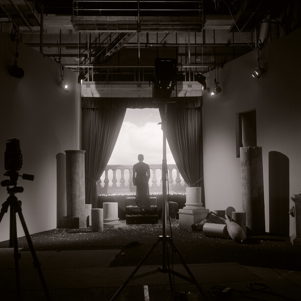 Studio setup with tripod-mounted camera in foreground, silhouetted figure standing on balcony framed by curtains and architectural columns.