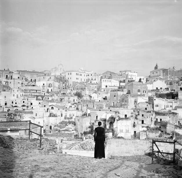 Woman in black dress standing on rooftop overlooking densely built hillside village with white-washed buildings.