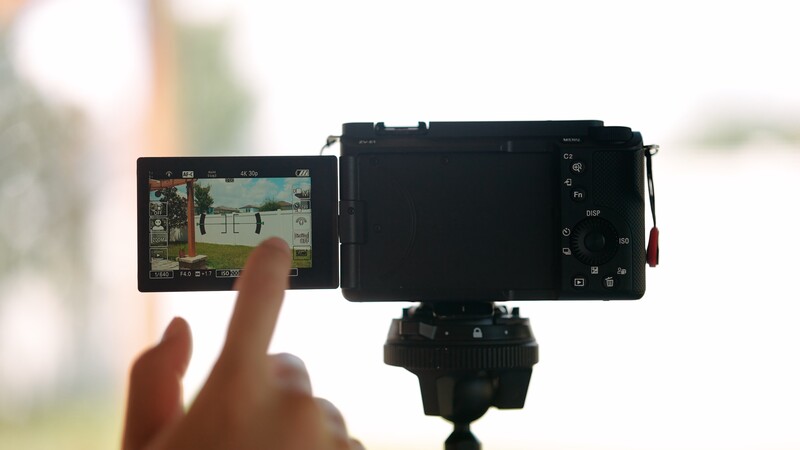 Hand touching the articulating touchscreen of a mirrorless camera mounted on a tripod, displaying a video preview of an outdoor scene.