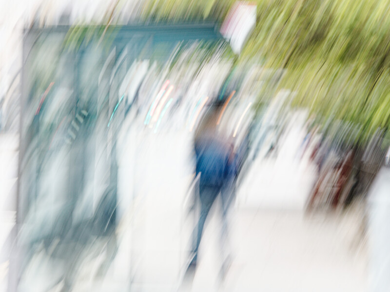 Blurred motion photograph of people walking on a street with green foliage visible.