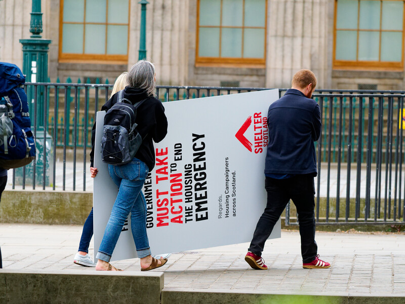 Two people carrying a large white poster board with red text and arrow graphic in front of a neoclassical building with columns.