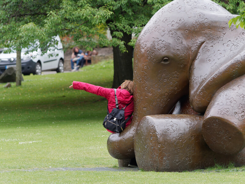 Child in bright pink jacket playfully interacting with a large bronze sculptural hand in a park setting.