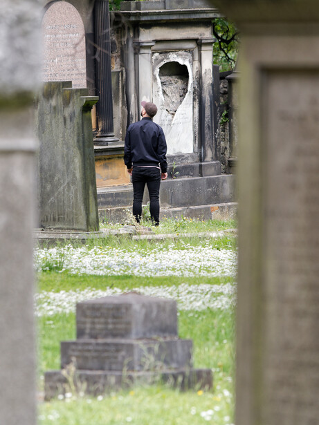 Person in dark jacket walking through cemetery between stone grave markers and mausoleum structures.