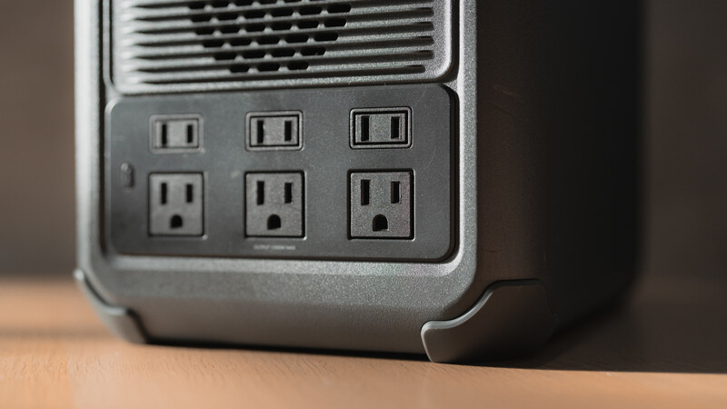 Close-up of a gray power strip mounted on a wooden surface, featuring six electrical outlets arranged in two rows and a ventilation grille above.