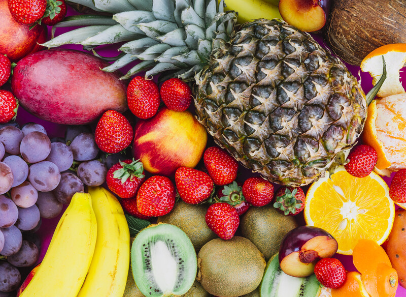 Overhead arrangement of fresh fruits including pineapple, strawberries, bananas, kiwi, grapes, and citrus on wooden surface.