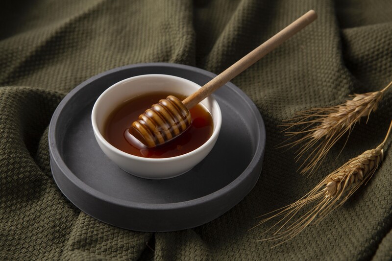 Honey drizzler resting in a white bowl filled with golden honey, placed on a dark gray plate atop olive green fabric with wheat stalks.