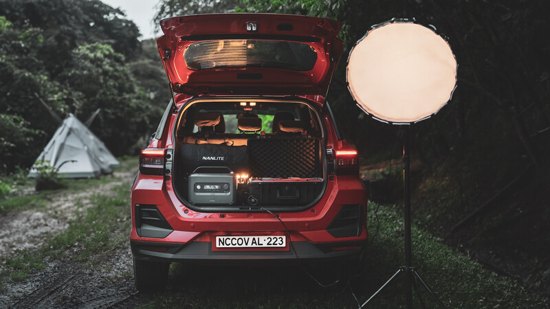 Red SUV with open trunk displaying camping gear at night, illuminated by a large circular reflector, with tent visible in background.