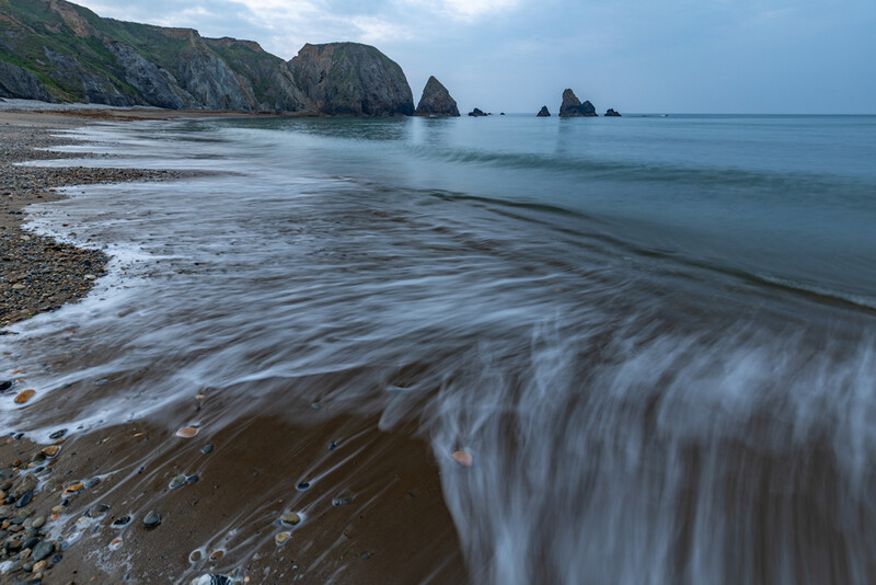 Seascape with long-exposure flowing water, rocky sea stacks, and overcast sky.