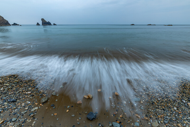 Long exposure seascape showing silky water motion flowing across a pebble beach at dusk.