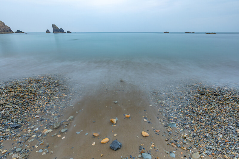 Long exposure seascape with smooth water, rocky beach, and distant sea stacks under overcast sky.