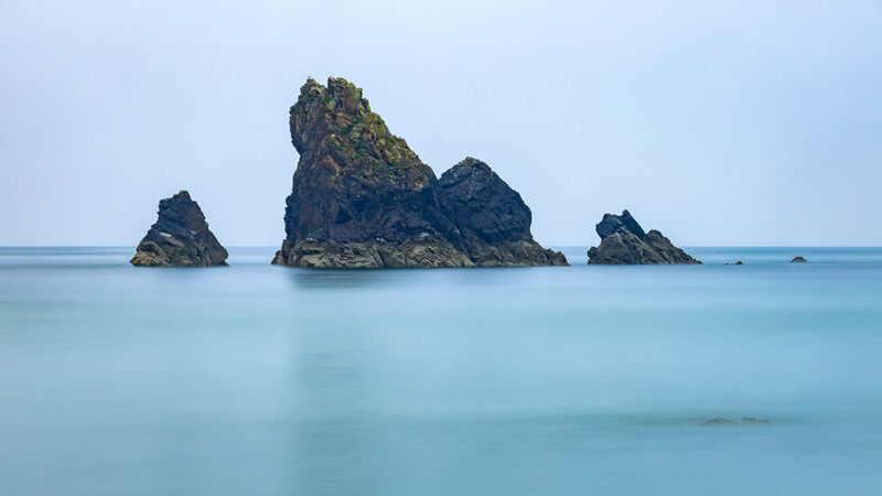 Three dark rocky islands with moss-covered peaks rising from calm turquoise water under clear sky.
