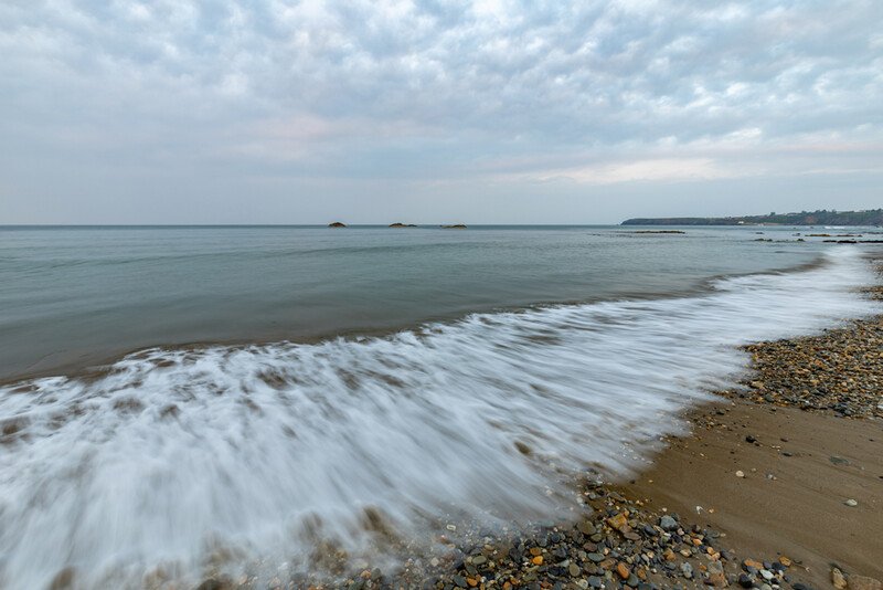 Waves roll onto a pebble and sand beach under an overcast sky, with a calm sea stretching toward a distant shoreline.
