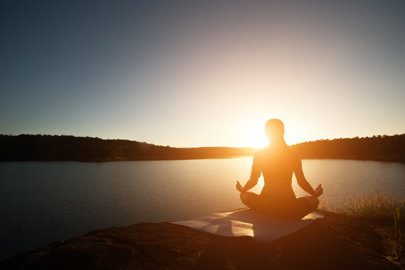 Silhouetted figure in meditation pose on a rock by a calm lake at sunset.