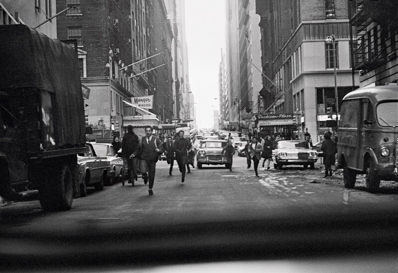 Busy urban street lined with tall buildings, pedestrians and vintage vehicles, shot from inside a vehicle looking forward.