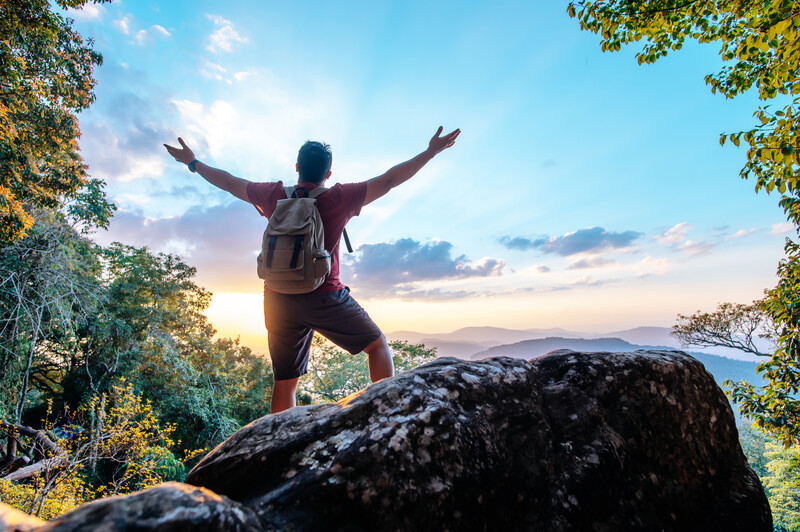 Hiker with arms raised standing on a boulder at sunset with mountains and water visible in the distance.