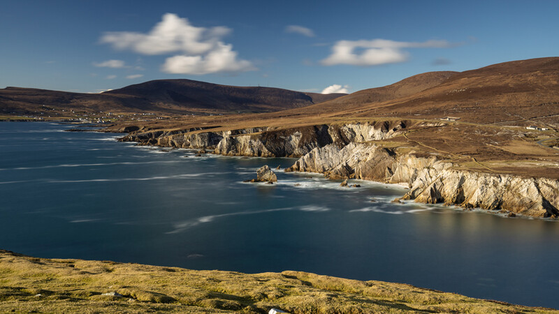 Dramatic coastal cliffs with turquoise water and rolling brown hills under partly cloudy sky.