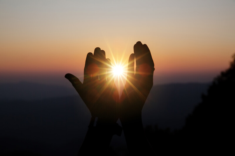 Silhouetted hands cupping a bright sun at golden hour against a dusky sky.