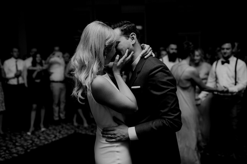 Newlyweds kiss on a darkened dance floor surrounded by wedding guests in formal attire.