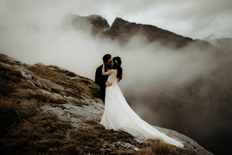 Groom in dark suit lifting bride in flowing white gown on rocky mountainside shrouded in mist.