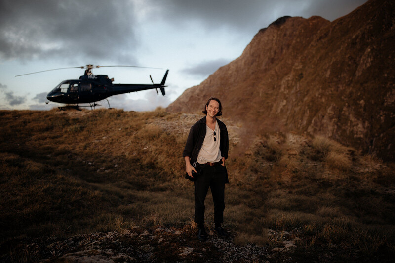 Man in dark blazer standing in barren landscape with black helicopter in background.