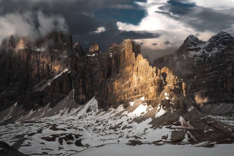 Dramatic mountain peaks with golden sunlit rock formations rising above snowy terrain under stormy skies.