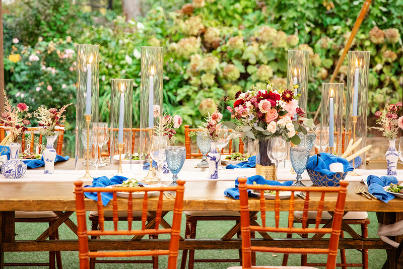 Outdoor garden wedding reception table decorated with tall glass vases, red and cream flowers, and blue napkins on wooden chairs.