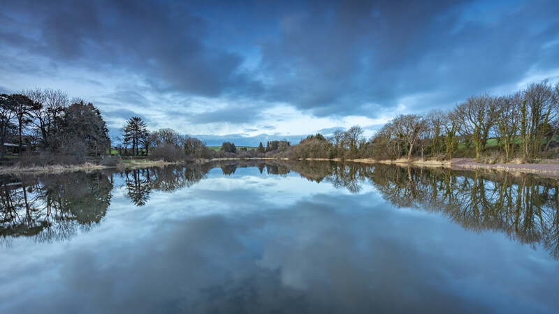 Calm lake with mirror-like reflections of bare trees and dramatic blue sky with clouds.