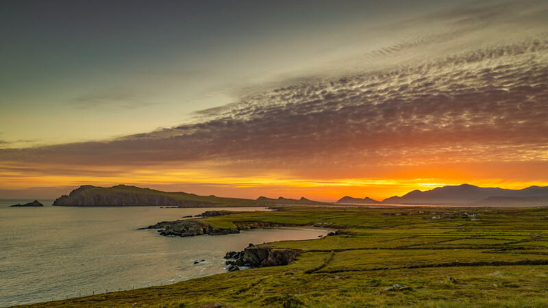 Dramatic golden sunrise over a coastal landscape with green fields, rocky outcrops, and distant mountains across the water.