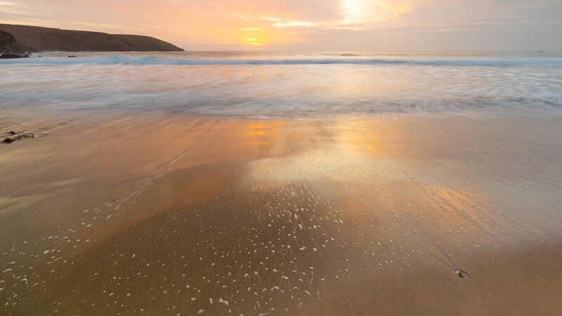 Golden sunset reflecting across wet sand and shallow water at a beach with headland in the distance.