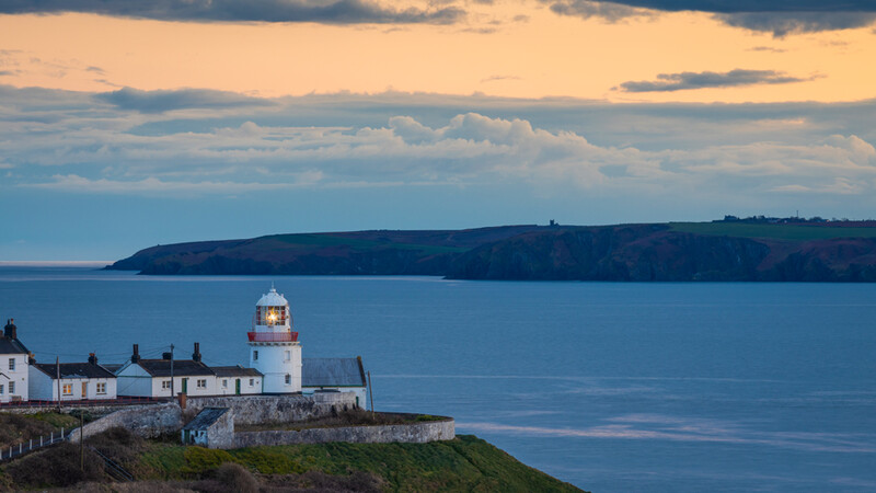 White lighthouse with stone buildings on a grassy headland overlooking calm blue water at sunset.