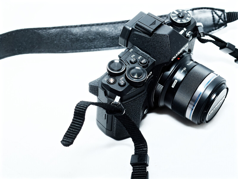 Black mirrorless camera with attached lens mounted on a tripod with strap against white background.